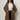 A woman with curly hair and glasses stands against a plain background, wearing the Sanctuary Clothing hometown jacket lynx extended sizing over a white button-up shirt, black satin pants, and keeps her hands in the coat pockets.