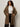 A woman with curly hair and glasses stands against a plain background, wearing the Sanctuary Clothing hometown jacket lynx extended sizing over a white button-up shirt, black satin pants, and keeps her hands in the coat pockets.