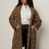 A woman with curly hair and glasses stands against a plain background, wearing the Sanctuary Clothing hometown jacket lynx extended sizing over a white button-up shirt, black satin pants, and keeps her hands in the coat pockets.