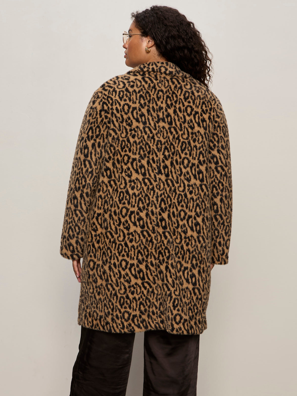 A person with curly hair and glasses is seen from behind, modeling the Sanctuary Clothing hometown jacket lynx extended sizing€�a knee-length, brown and black leopard-print coat€�against a plain light background.