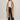 A woman with straight brown hair and glasses stands against a plain light wall, wearing the Sanctuary Clothing "live in fur coat warm camel" over a white tee, high-waisted dark blue jeans with buttons, and black shoes.
