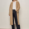 A woman with straight brown hair and glasses stands against a plain light wall, wearing the Sanctuary Clothing "live in fur coat warm camel" over a white tee, high-waisted dark blue jeans with buttons, and black shoes.