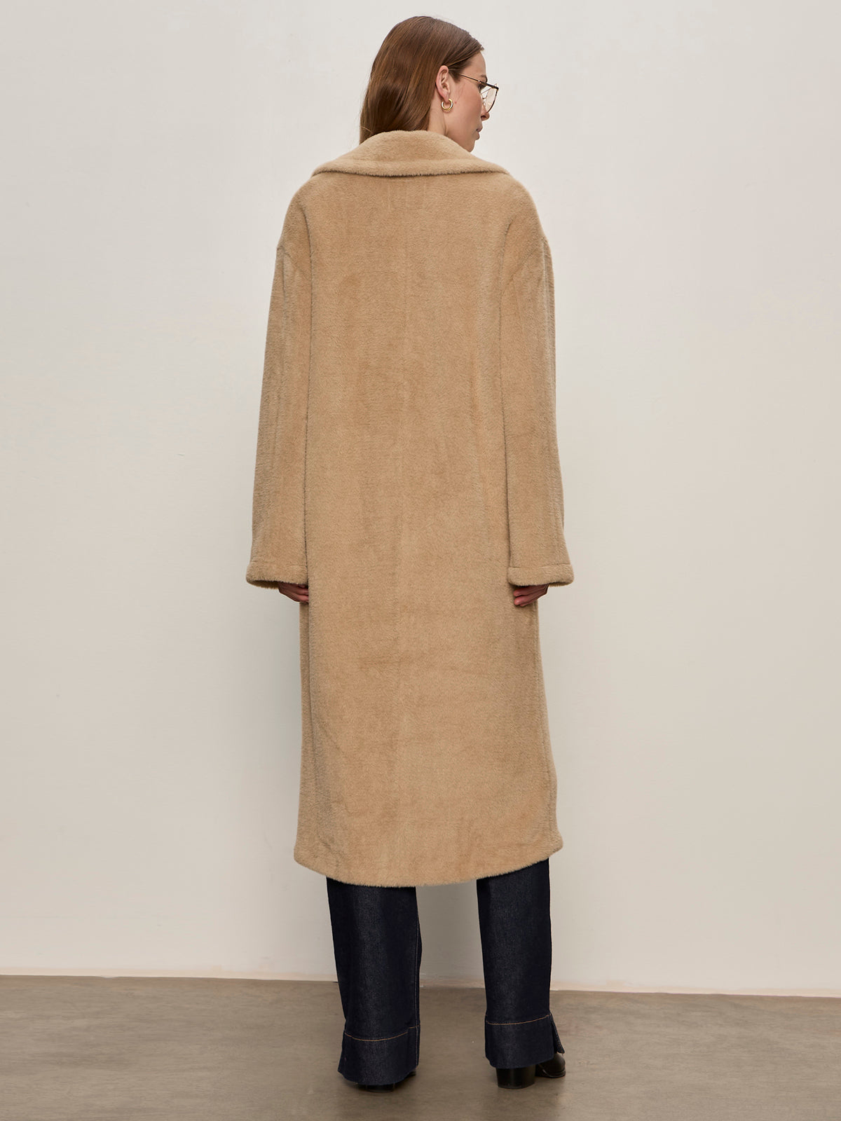 A person with long brown hair and glasses wears the Sanctuary Clothing "live in fur coat warm camel," paired with dark wide-leg jeans and black shoes, standing against a plain off-white wall.