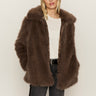 A woman with straight blonde hair wears the Sanctuary Clothing carmen fur coat in morel over a white top and dark jeans, standing against a plain light background.