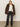 A woman poses against a plain background wearing the Sanctuary Clothing carmen fur coat morel over a white sweater, dark high-waisted jeans, and black boots, with one hand in her pocket and the other holding her coat open.
