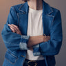 A person wearing Sanctuary Clothing's "sculpted denim blazer best in show" over a white shirt, arms crossed. Gold hoop earrings, a gold bangle, and the blazer's rolled-up sleeves accentuate dark pants with a belt against a soft gradient background.