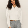 A woman with curly hair models the Sanctuary Clothing cropped paper boy jacket in bleached white, featuring a chest pocket, against a plain beige background.