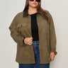 A woman with long brown hair wears Sanctuary Clothing's barn jacket in burnt olive (extended sizing) over a black top and blue jeans, paired with dark sunglasses, standing against a plain light background and looking at the camera.