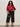 A woman with curly hair wears the Sanctuary Clothing satin track jacket in crimson (extended sizing) with white sleeve stripes, wide-leg dark pants, black heeled sandals, and a studded black handbag. She stands against a plain light wall.