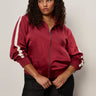 A woman with curly hair models the Sanctuary Clothing satin track jacket in crimson (extended sizing), featuring white sleeve stripes. She pairs it with dark pants and a black studded purse, posing against a light background.