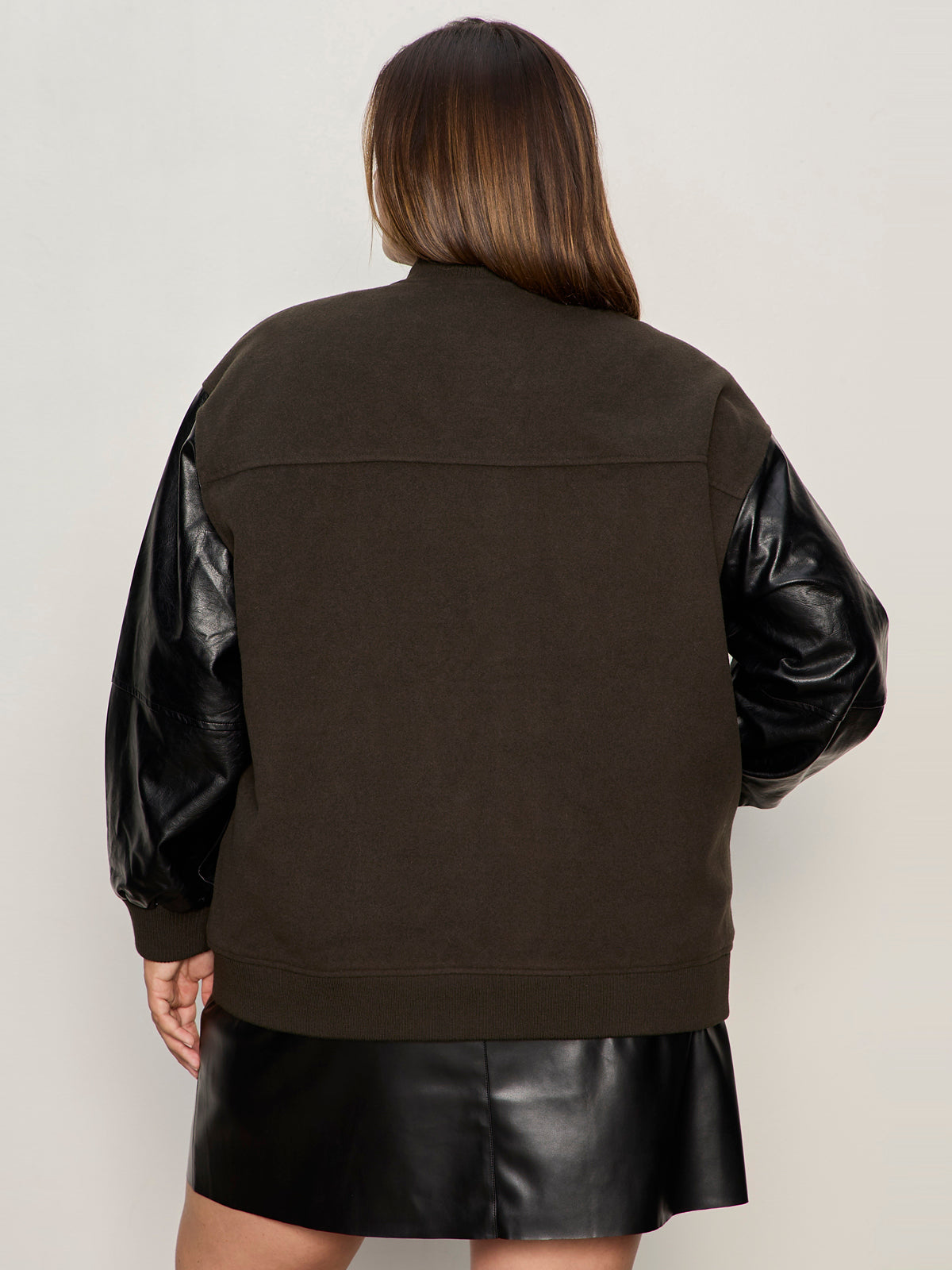 A woman with long brown hair, facing away, wears the Sanctuary Clothing club varsity jacket in midnight olive (extended sizing) featuring black faux leather sleeves and a matching skirt, against a plain light-colored background.