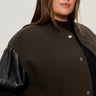 A woman with long brown hair wears the Sanctuary Clothing club varsity jacket in midnight olive (extended sizing), featuring black leather sleeves and silver buttons, standing against a neutral background.