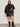 A woman stands against a plain light wall wearing Sanctuary Clothing's club varsity jacket in midnight olive (extended sizing), paired with a black sweater, black leather skirt, and black boots. She faces forward, holding her jacket with one hand.