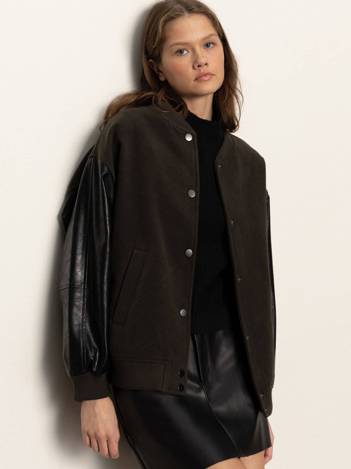 A young woman with long brown hair wears the Sanctuary Clothing club varsity jacket in midnight olive, featuring leather sleeves. She stands against a plain light background, facing the camera with a neutral expression.