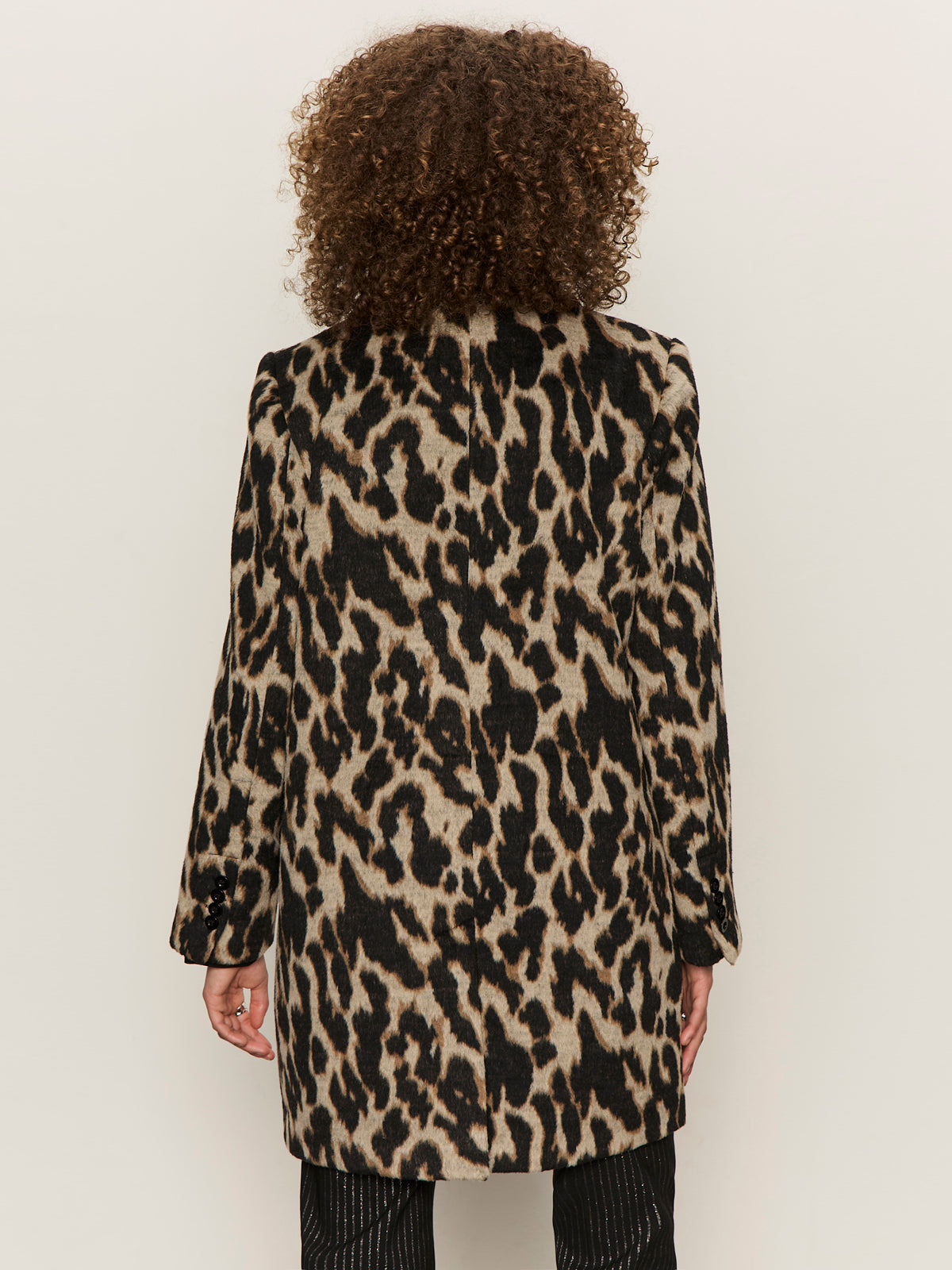 Seen from behind, a person with curly hair wears the Sanctuary Clothing downtown jacket leopard mirage€�featuring black, brown, and beige leopard print€�and black pants against a plain light background.