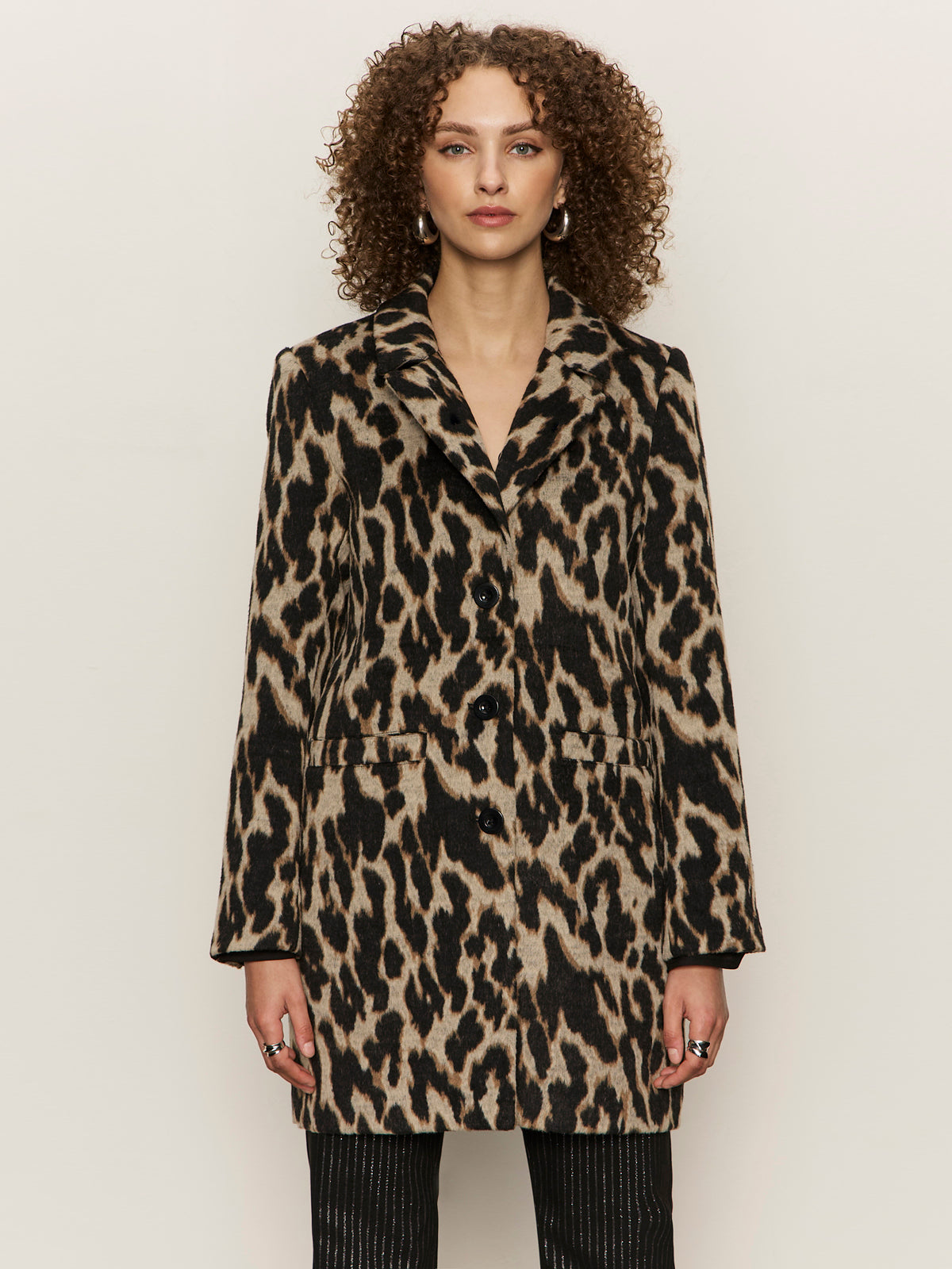 Someone with curly hair wears the Sanctuary Clothing downtown jacket leopard mirage€�a long, buttoned coat with a bold leopard print€�paired with black striped pants, standing against a plain light background.