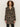 Someone with curly hair wears the Sanctuary Clothing downtown jacket leopard mirage€�a long, buttoned coat with a bold leopard print€�paired with black striped pants, standing against a plain light background.