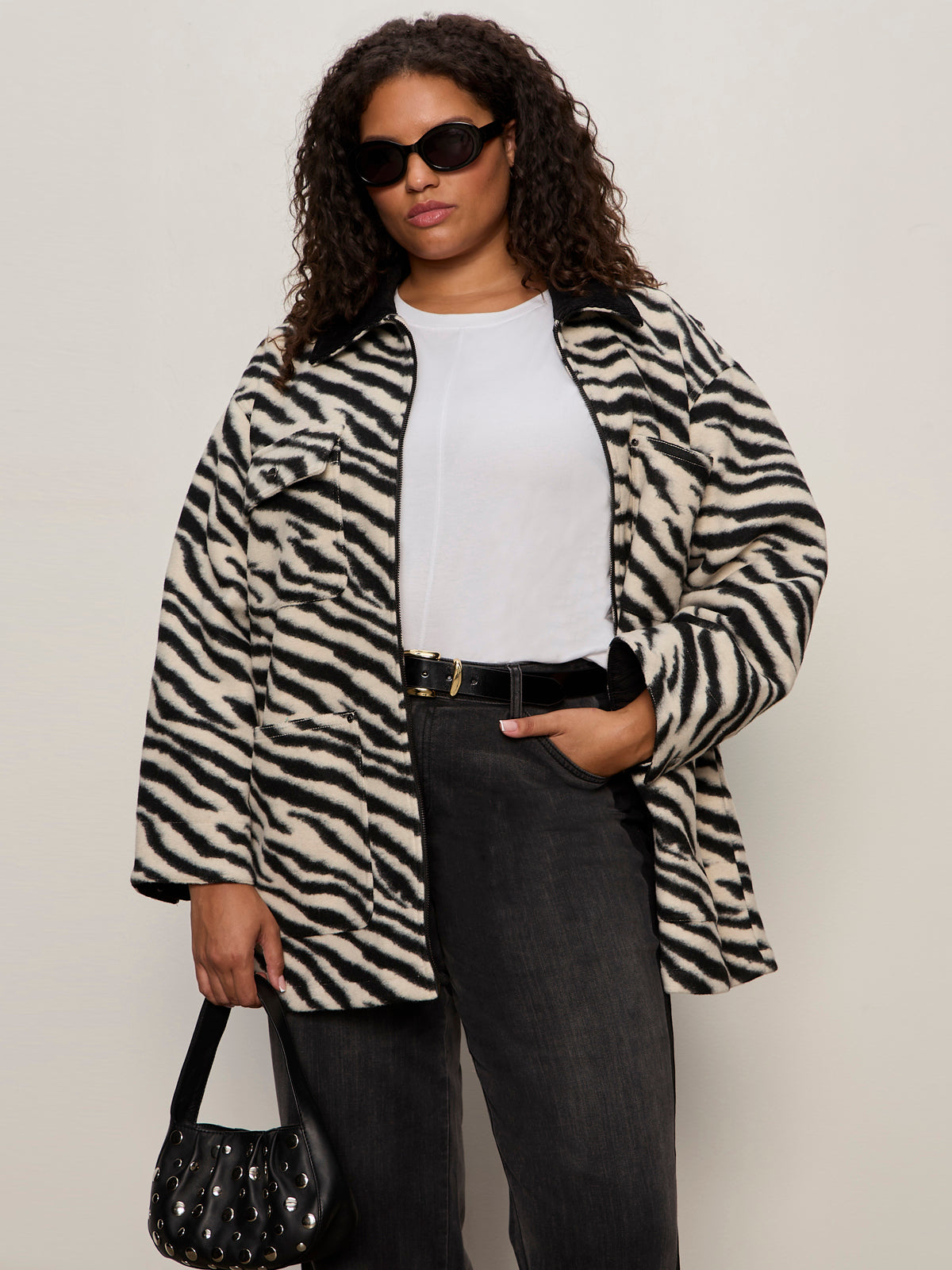 A woman wears the Sanctuary Clothing soiree barn jacket striking zebra (extended sizing) over a white tee and dark jeans, accessorized with black sunglasses, a belt, and handbag, standing against a plain light background.