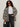 A woman wears the Sanctuary Clothing soiree barn jacket striking zebra (extended sizing) over a white tee and dark jeans, accessorized with black sunglasses, a belt, and handbag, standing against a plain light background.