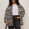 A woman wears the Sanctuary Clothing soiree barn jacket striking zebra (extended sizing) over a white tee and dark jeans, accessorized with black sunglasses, a belt, and handbag, standing against a plain light background.