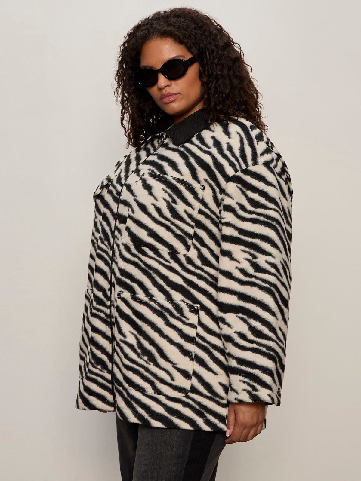 A woman with curly hair and sunglasses wears the Sanctuary Clothing soiree barn jacket striking zebra extended sizing, featuring a black collar, against a plain light background.