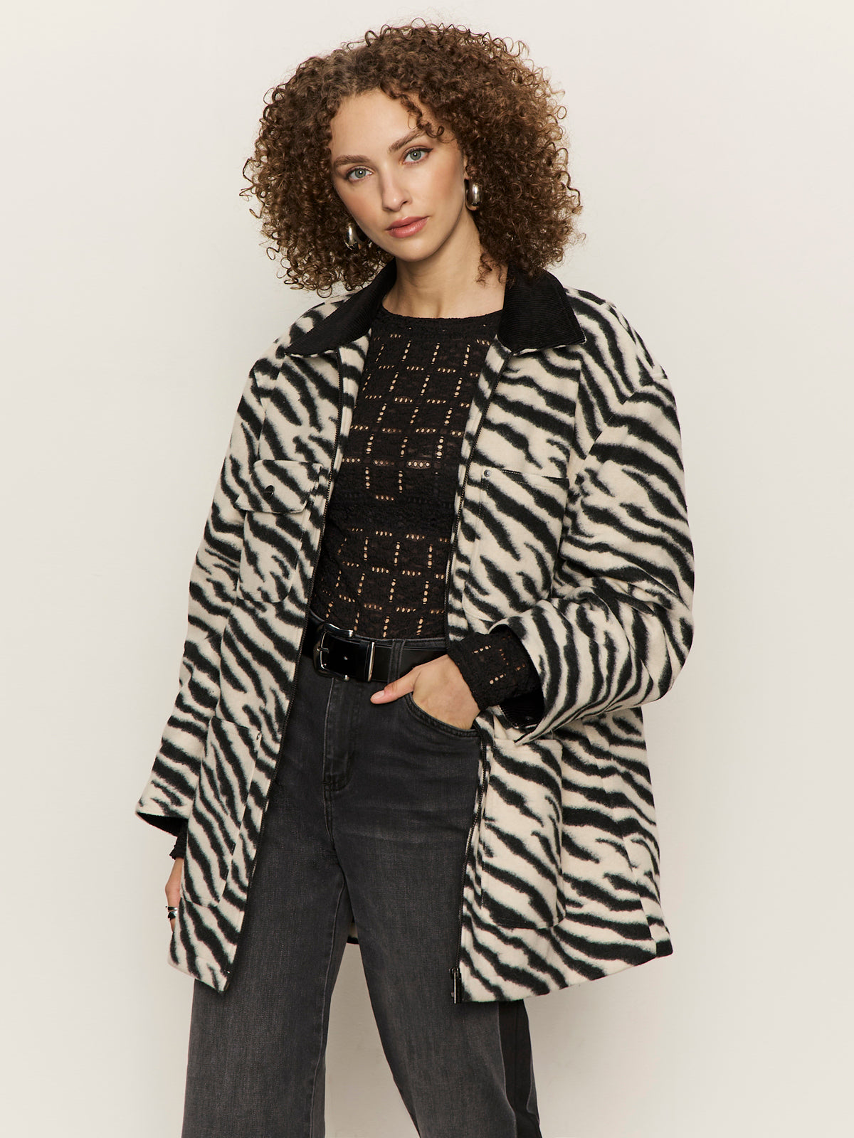 A woman with curly hair wears a black and white zebra-print coat over a black patterned sweater and dark jeans, standing against a plain light background.