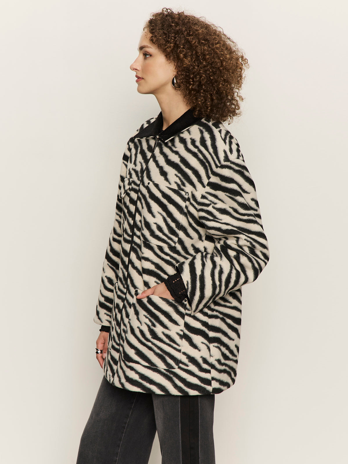A woman with curly hair wears a black and white zebra-print coat over black pants, standing in profile against a plain light background, with one hand in her coat pocket.