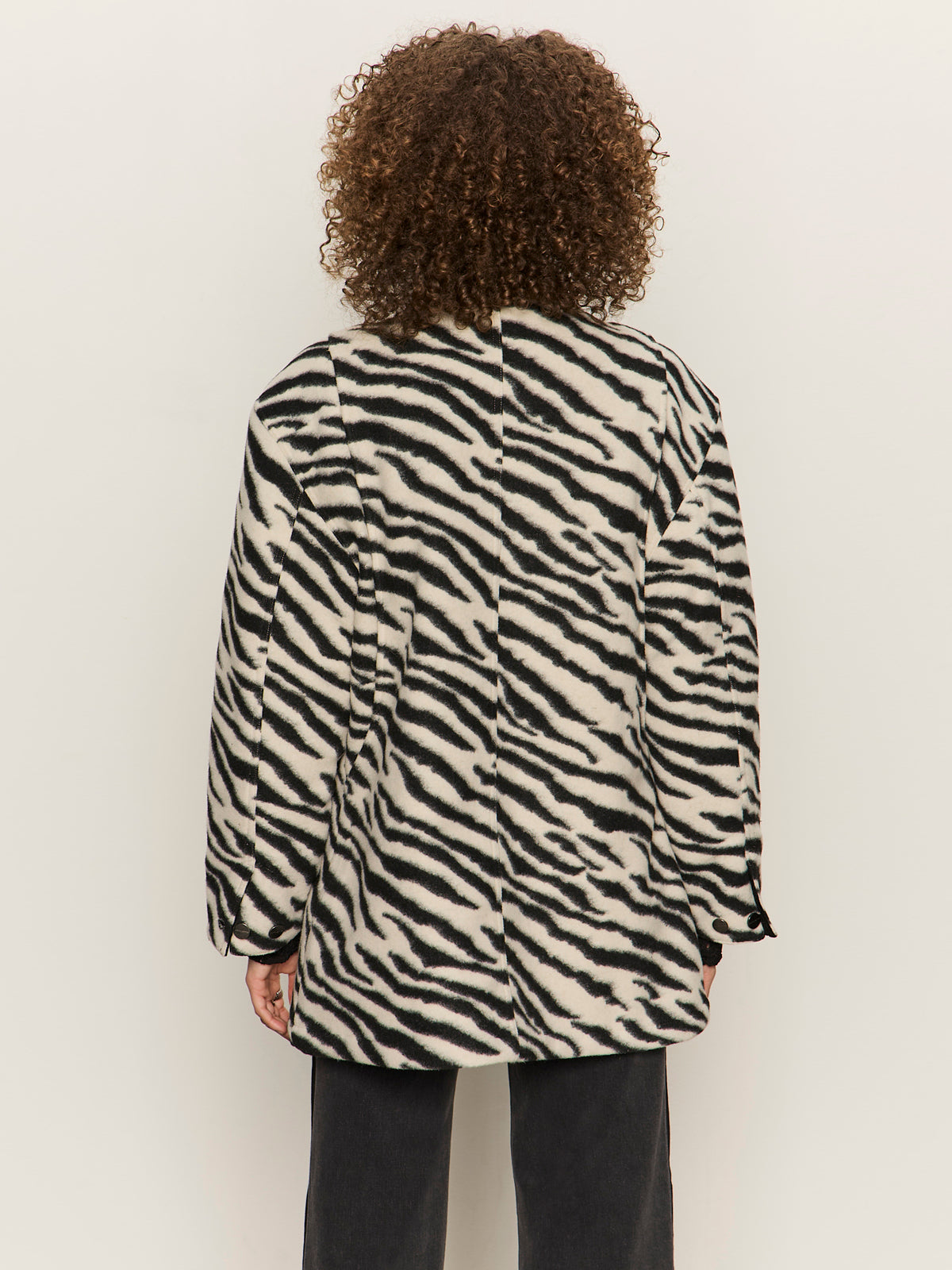 A person with curly hair, seen from behind, wearing a long-sleeve black and white zebra print coat and black pants, standing against a plain light background.