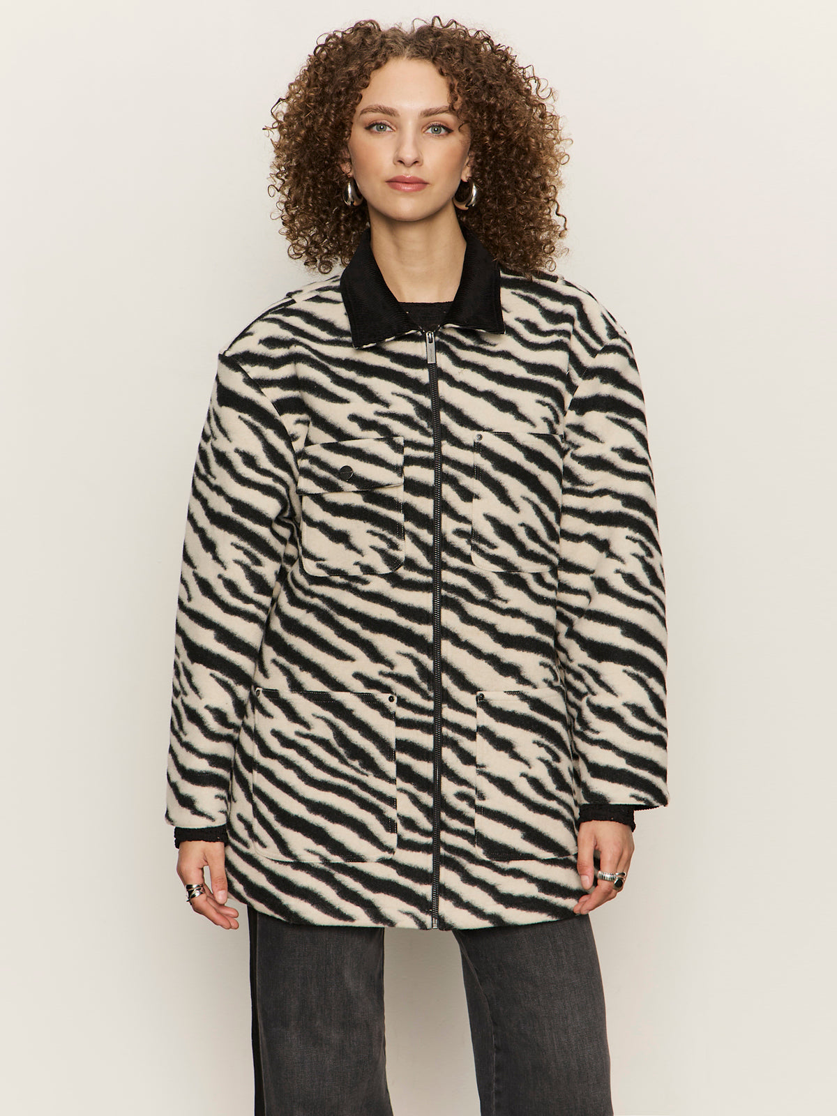 A woman with curly hair models the Sanctuary Clothing Soiree Barn Jacket Striking Zebra, featuring a black collar, paired with dark pants and accessories, standing against a plain light background.