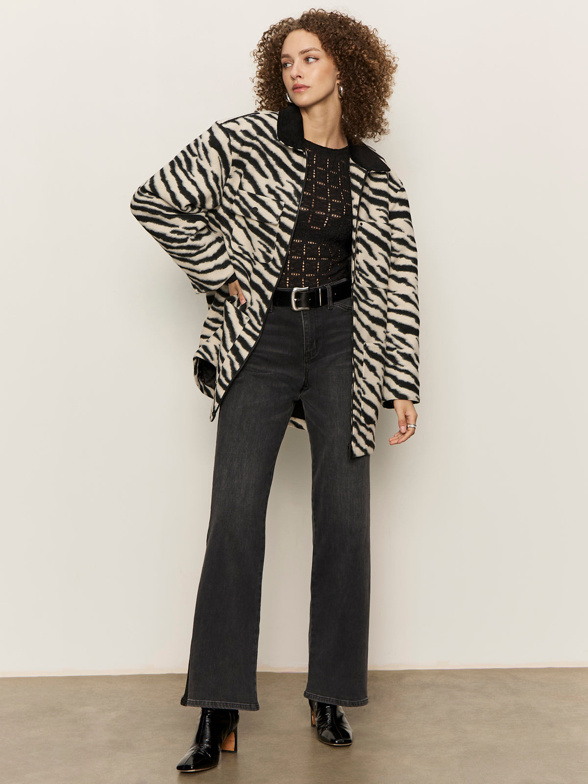 Wearing Sanctuary Clothing's soiree barn jacket striking zebra, a person with curly hair pairs it with a black patterned top, black belt, dark gray jeans, and black heeled boots against a plain light-colored wall.