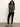 A woman with long curly hair stands against a plain light wall, wearing Sanctuary Clothing™s runway legging in black, a dark gray long-sleeve top, and black ankle-strap flats. She has her arms crossed and looks at the camera.