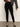 A person models the Sanctuary Clothing runway legging black, paired with a loose dark gray long-sleeve top and black ankle-strap shoes, posed with one hand on their hip against a plain light background, head out of frame.