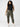 A woman with curly hair stands against a simple background, wearing a black sleeveless top, Rebel Pant Hiker Camo from the Inclusive Collection by Sanctuary Clothing, and white sneakers. She has her hands in her pockets and gazes directly at the camera.