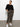 A woman stands against a plain wall, wearing Sanctuary Clothing's rebel pant hiker camo from the inclusive collection, paired with a loose black sweater and white sneakers, her hands in her pockets as she looks to the side.