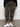 A person wears Sanctuary Clothing's rebel pant hiker camo from the Inclusive Collection, paired with a black sweater and white sneakers, standing indoors on a gray floor against a plain white wall.