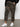 Wearing Sanctuary Clothing's rebel pant hiker camo from the inclusive collection, a person stands against a plain background with one hand in their pocket, paired with a black sweater and white sneakers.