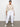 A woman stands against a plain white wall, hands in her pockets, wearing the rebel pant in brilliant white from Sanctuary Clothing™s Inclusive Collection, paired with a beige knit sweater and white sneakers as she looks confidently at the camera.