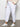 A person stands indoors wearing Sanctuary Clothing's rebel pant brilliant white from the inclusive collection, paired with a cream knit sweater and white sneakers with light-colored accents.