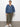 A woman stands against a white wall wearing Sanctuary Clothing™s rebel pant hiker green from the inclusive collection, paired with a loose denim blouse and white sneakers. One hand in her pocket, she maintains a neutral expression.