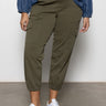 A person in Sanctuary Clothing's rebel pant hiker green (inclusive collection), a blue long-sleeve top, and white sneakers stands on a gray floor against a white wall. The photo is cropped just below the shoulders.