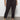 Wearing the rebel pant obsidian from Sanctuary Clothing's inclusive collection, paired with a green camo tee and white sneakers, a person stands against a plain wall, hands in pockets.