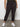 Wearing the rebel pant obsidian from Sanctuary Clothing's inclusive collection, paired with a green camo tee and white sneakers, a person stands against a plain wall, hands in pockets.