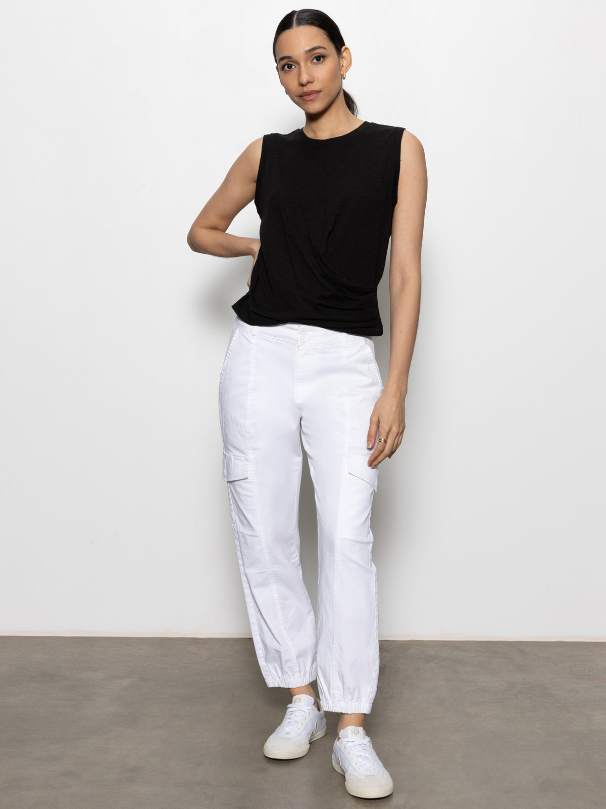 A woman stands against a white wall wearing Sanctuary Clothing™s rebel pant in brilliant white, paired with a sleeveless black top and white sneakers. Her hair is tied back and one hand rests on her hip.