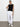 A woman stands against a white wall wearing Sanctuary Clothing™s rebel pant in brilliant white, paired with a sleeveless black top and white sneakers. Her hair is tied back and one hand rests on her hip.