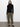 A person with long, straight dark hair and a neutral expression stands against a plain white wall, wearing a black turtleneck, black woven shoes, and the Sanctuary Clothing rebel pant in hiker green.