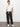 A woman stands against a white wall, wearing a white short-sleeve knotted tee, Sanctuary Clothing's rebel pant in obsidian, and white sneakers, with her hands in her pockets and a relaxed expression.
