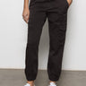 Wearing a white t-shirt, white sneakers, and Sanctuary Clothing's rebel pant obsidian cargo pants with elastic cuffs, a person stands against a plain, light-colored wall with hands in pockets.