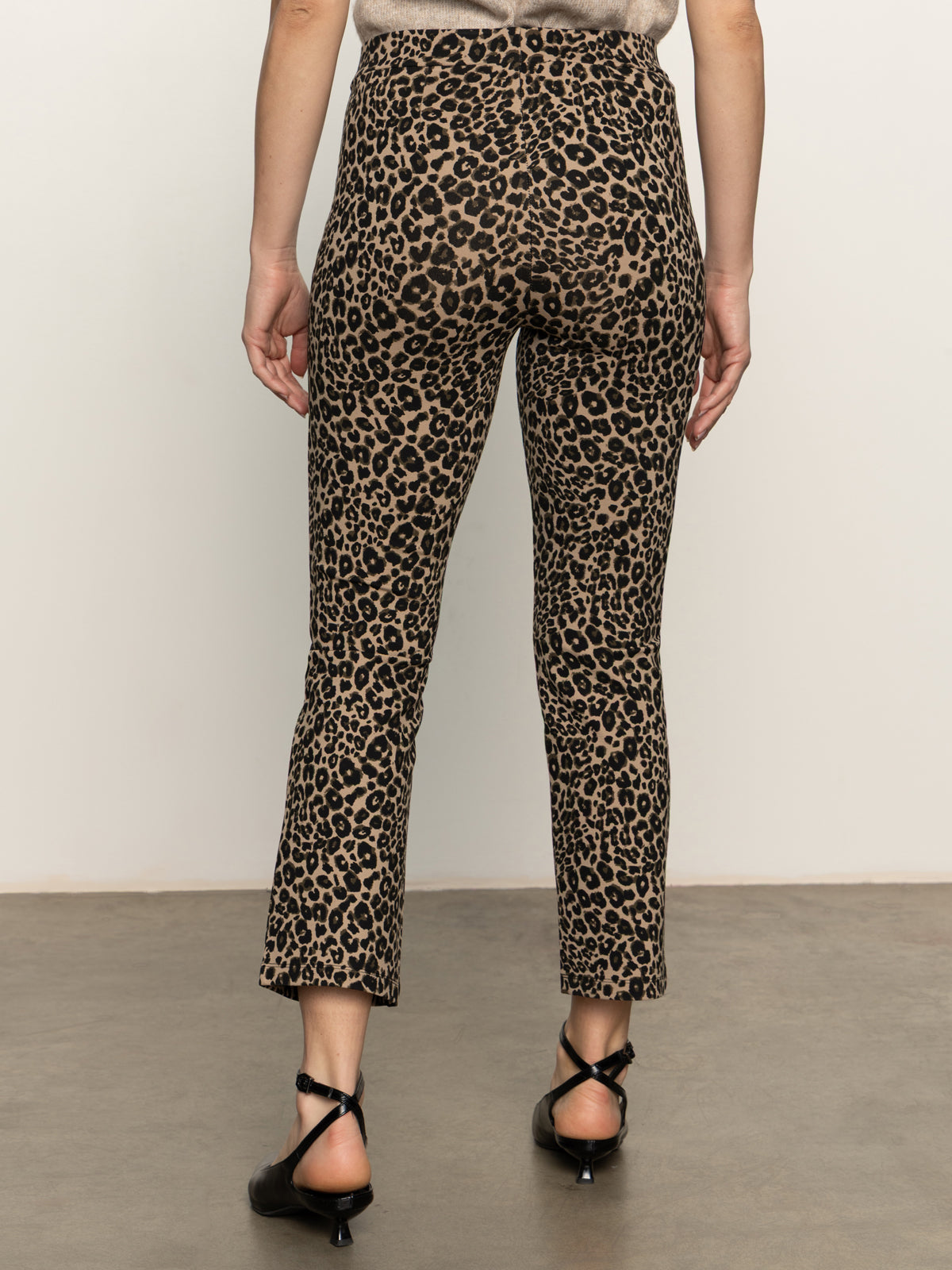 A person stands facing away from the camera, wearing Sanctuary Clothing’s carnaby kick crop leggings in French Kitty print with black strappy low-heeled shoes. The background is neutral and plain.