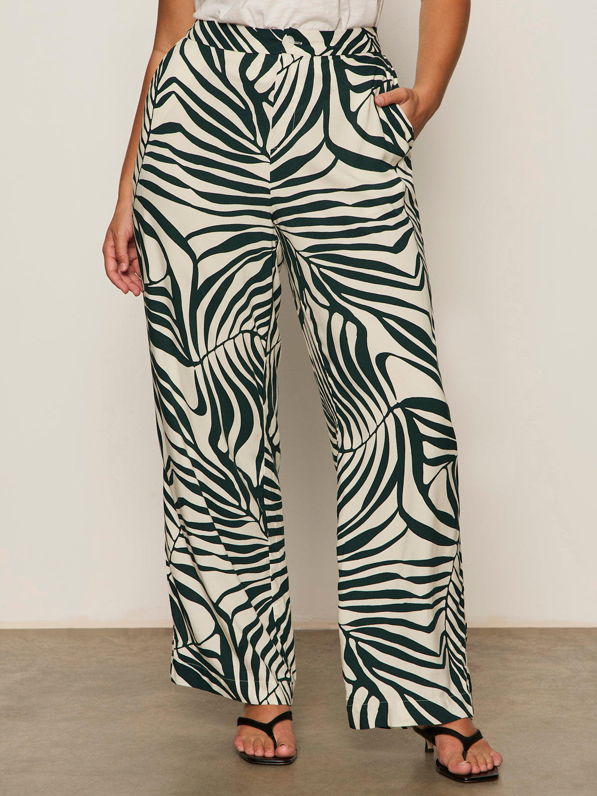 Someone wearing Sanctuary Clothing’s the soft trouser green oasis extended sizing, styled with black sandals and a tucked-in white top, stands against a plain background.