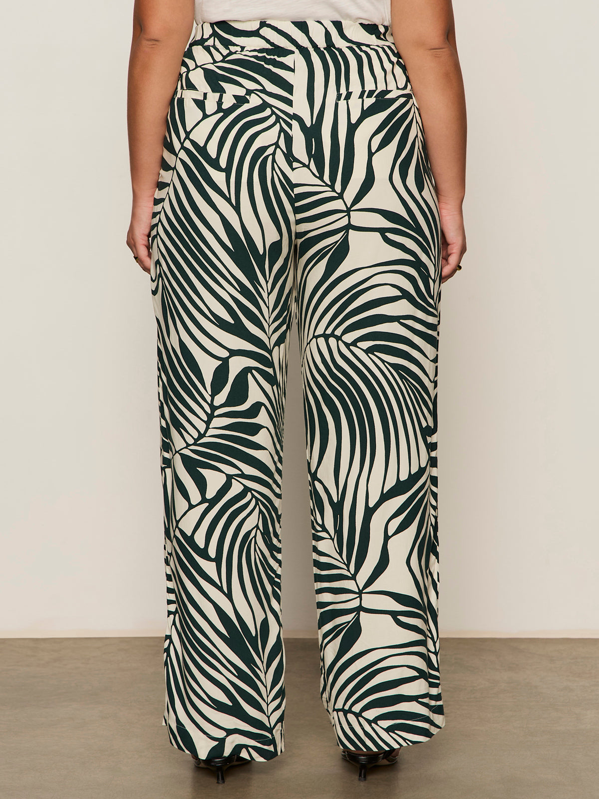 A person stands facing away, wearing the soft trouser green oasis from Sanctuary Clothing—high-waisted, wide-leg, loose-fitting pants in extended sizing. The background is plain and neutral.